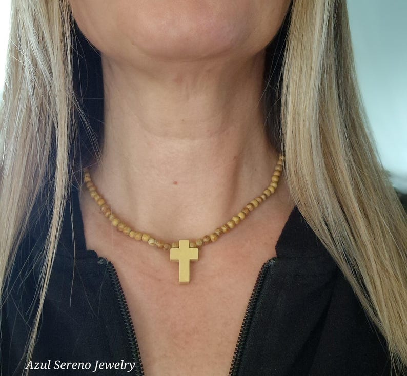 May include: A necklace featuring a series of small, round, light brown wooden beads strung together. A smooth, solid gold-colored cross pendant hangs from the center of the bead strand. The necklace rests against the neck, showcasing the contrast between the natural wood texture and the metallic finish of the cross.