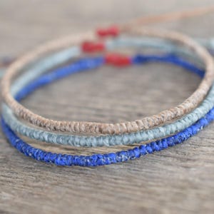 May include: Three handmade bracelets in neutral tones. The bracelets are stacked, with a light brown, a light blue, and a vibrant blue. Each bracelet has a knotted design and red accents. The bracelets are on a wooden surface.