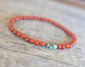 Puede incluir: Un brazalete de piedras rojas con un solo acento de cuentas turquesa. El brazalete está hecho de pequeñas cuentas redondas y está enhebrado en elástico.