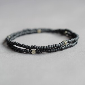 May include: A double-strand bracelet composed of small, matte black beads. The bracelet features small, square, metallic accents. The jewelry is displayed on a neutral gray background, highlighting the bracelet's dark color and minimalist design.