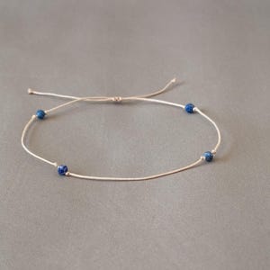 May include: A delicate bracelet featuring a thin, light-colored cord with three small, round, deep blue beads spaced evenly along the strand. The bracelet includes an adjustable sliding knot closure with two loose cord ends extending outward.