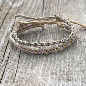 Peut inclure: Bracelets tressés aux tons neutres de beige, de tan et de lavande. Les bracelets sont faits de cordon tissé et ont des fermetures réglables. Les bracelets sont présentés sur une surface en bois vieilli.