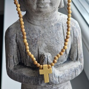 May include: A necklace featuring small, round, light brown wooden beads and a central light yellow wooden cross pendant. The necklace is draped over a grey stone statue of a figure with hands in a prayer position. The text Azul Sereno Jewelry is visible.