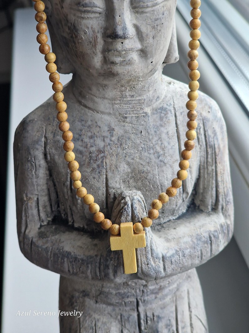 May include: A necklace featuring small, round, light brown wooden beads and a central light yellow wooden cross pendant. The necklace is draped over a grey stone statue of a figure with hands in a prayer position. The text Azul Sereno Jewelry is visible.