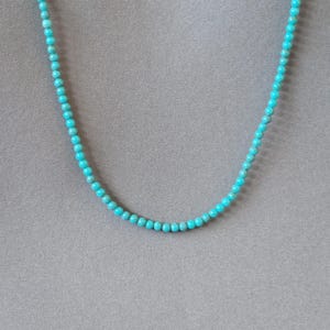 Peut inclure: Un collier délicat composé de petites perles rondes turquoise. Les perles sont enfilées pour former un seul brin, créant un bijou simple mais élégant. Le collier est présenté sur un fond gris neutre.