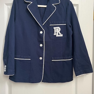 May include: Navy blue jacket with white trim and a white embroidered "R" on the pocket. The jacket has a notched collar, three buttons on the sleeve cuffs, and two front pockets. The jacket is hanging on a white hanger.