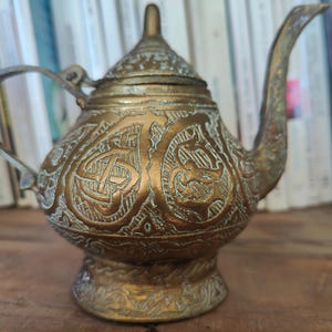 Può includere: Una teiera in metallo color oro con intricati disegni incisi. La teiera ha un corpo arrotondato e un beccuccio con un manico curvo.