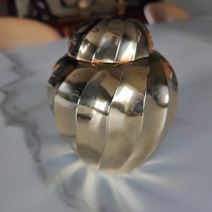 Könnte beinhalten: Eine goldene dekorative Metalldose mit geripptem Design und Deckel. Die Dose steht auf einer weißen Oberfläche.