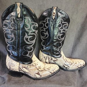 May include: A pair of cowboy boots with a black leather upper and a snakeskin-patterned foot. The boots have decorative stitching in a swirling pattern and a pointed toe. The shaft of the boots is approximately 12 inches high.