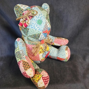 May include: A stuffed teddy bear constructed from various patchwork fabric squares in shades of blue, red, yellow, and green. The bear features a green button eye, a jute twine bow around its neck, and decorative fabric flowers with pink beads.
