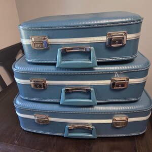 May include: A stack of three blue suitcases with a textured finish, each featuring a white horizontal stripe, silver metal latches, and a matching blue plastic handle. The suitcases are arranged in descending order of size from top to bottom.