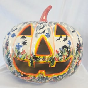 May include: A ceramic Halloween pumpkin with a carved face and triangular eye cutouts. The pumpkin is cream-colored with orange and yellow accents. It features painted bats, spiders, and floral patterns, creating a festive and decorative piece for the holiday.
