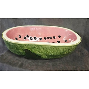 May include: An oval ceramic bowl shaped like a halved watermelon. The interior is painted a soft pink with scattered black teardrop-shaped seeds. The exterior features a textured green rind with a dark green leaf detail on the side.