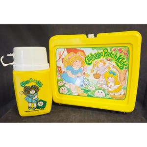 May include: A yellow Cabbage Patch Kids lunchbox and matching thermos. The lunchbox features a colorful illustration of Cabbage Patch Kids. The thermos has a white lid and handle. Both items have the Cabbage Patch Kids logo.