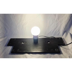 May include: A lit, round, white LED light bulb is screwed into a black base with a power cord. The base is rectangular with a unique shape and two holes. The background is a white sheet.