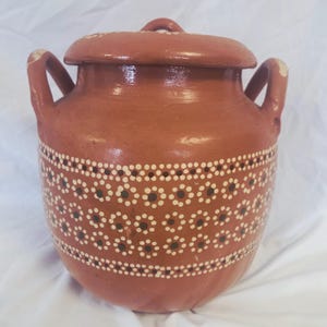May include: A terracotta-colored ceramic pot with a lid and two handles. The pot is decorated with a white and black dotted floral pattern around the middle. The lid has a small loop handle.
