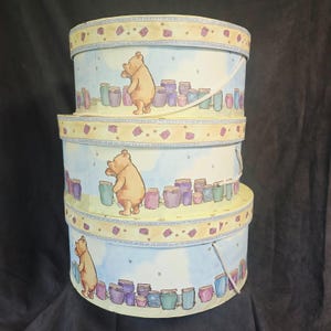 May include: A stack of three round, light blue storage boxes with lids. Each box features a repeating illustration of a yellow bear standing near multiple colorful honey pots. The lids have a yellow border with small purple square patterns.