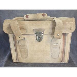 May include: A tan suede Ramrod camera bag with a top handle and a metal clasp closure. The bag has a rectangular shape with a flap, two zippers, and the brand name printed on the front.