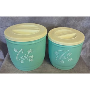 May include: Two turquoise canisters with off-white lids. The larger canister features the white text "Cobbee" surrounded by small flower icons, while the smaller canister features the white text "Tea" with similar flower icons.