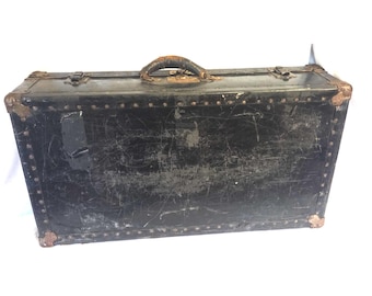 May include: A vintage black trunk with a worn, distressed appearance. The trunk features a leather handle, metal hardware, and corner protectors. Rivets line the edges, and the trunk shows signs of age and use.
