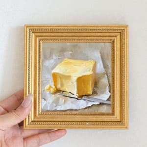 May include: A small, square, gold-framed painting of a butter sculpture. The butter is a block of yellow, sitting on white paper. A silver knife rests on the paper. The painting is on a white background.