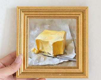 May include: A framed oil painting depicts a square block of yellow butter on white paper, with a silver knife. The painting is in a gold frame. The artwork is a still life, capturing the texture and color of the butter.