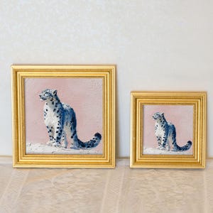 May include: Two framed paintings of snow leopards. The paintings feature a blue and white snow leopard against a pink background. The frames are gold. The paintings are of different sizes, with the larger one on the left and the smaller one on the right.