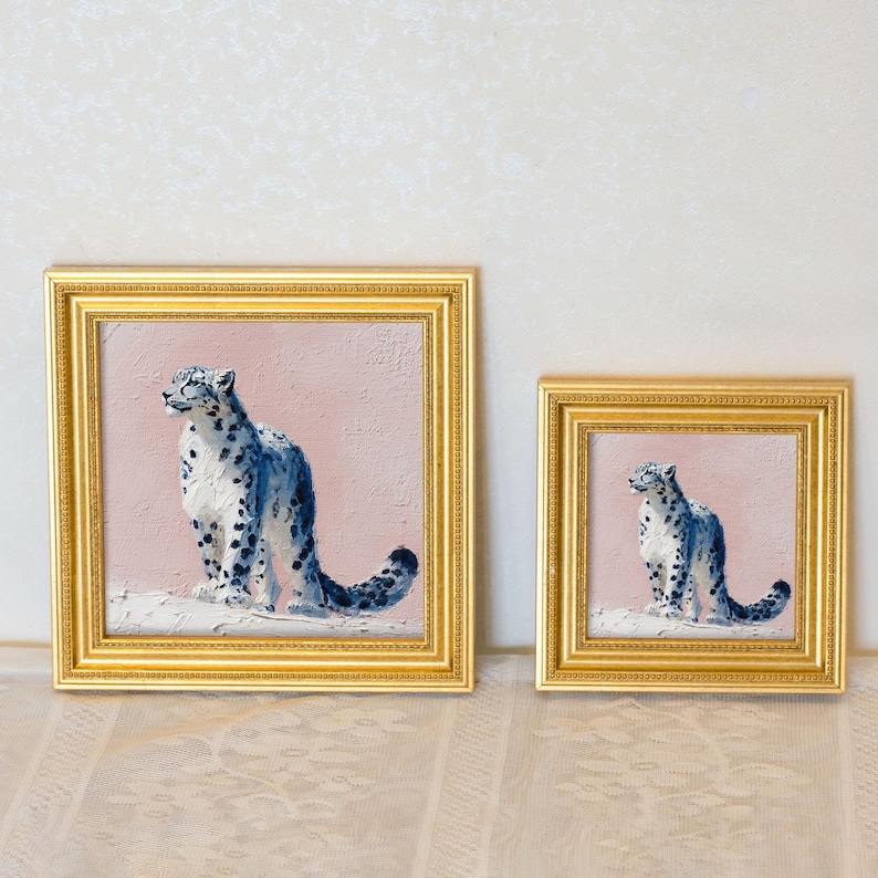 May include: Two framed paintings of snow leopards. The paintings feature a blue and white snow leopard against a pink background. The frames are gold. The paintings are of different sizes, with the larger one on the left and the smaller one on the right.