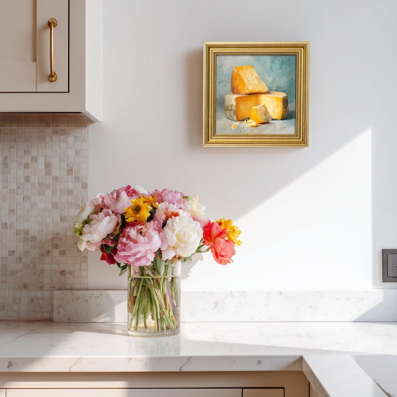 Peut inclure: Un tableau encadr&eacute; de morceaux de fromage est accroch&eacute; au-dessus d'un comptoir. Un bouquet de fleurs roses, jaunes et rouges dans un vase transparent est pos&eacute; sur le comptoir. Le tableau a un cadre dor&eacute; et est mont&eacute; sur un mur blanc.