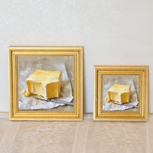 May include: Two framed oil paintings of butter. Each painting depicts a square of yellow butter on a piece of paper with a knife. The paintings are in gold frames. The butter is a light yellow color.