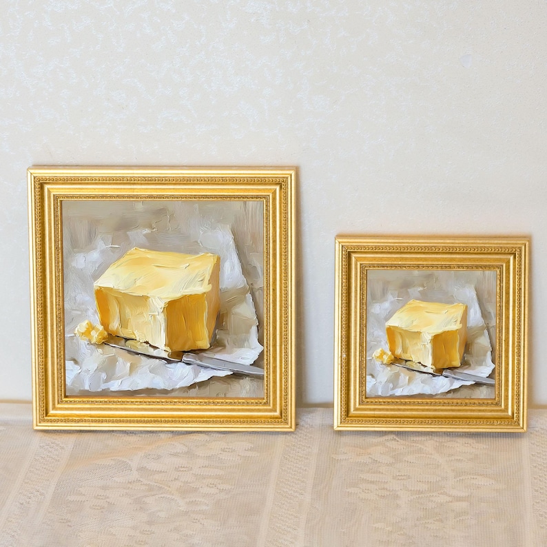 May include: Two framed oil paintings of butter. Each painting depicts a square of yellow butter on a piece of paper with a knife. The paintings are in gold frames. The butter is a light yellow color.