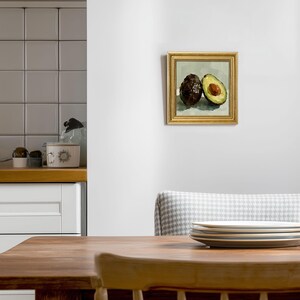 Op de afbeelding: Een ingelijst schilderij van twee avocado's. E&eacute;n hele, donkerbruine avocado staat naast een gehalveerde avocado, met een groene binnenkant en een bruine pit. Het schilderij heeft een gouden lijst en hangt aan een witte muur.
