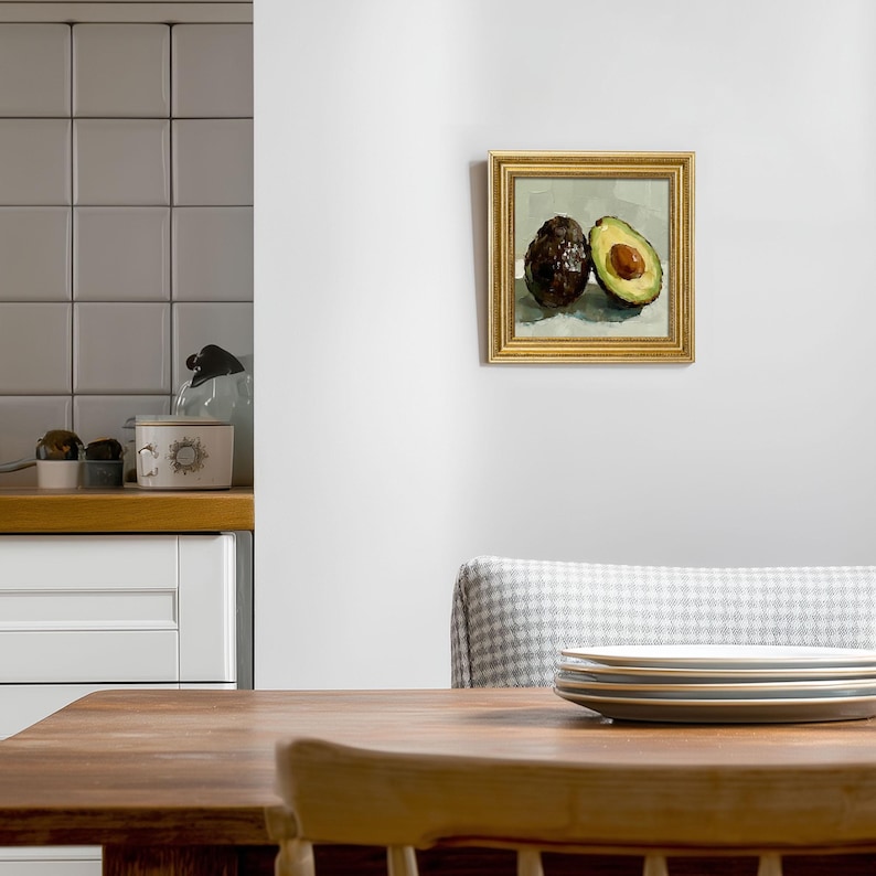 Op de afbeelding: Een ingelijst schilderij van twee avocado's. E&eacute;n hele, donkerbruine avocado staat naast een gehalveerde avocado, met een groene binnenkant en een bruine pit. Het schilderij heeft een gouden lijst en hangt aan een witte muur.