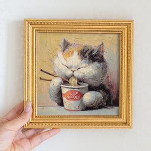 Op de afbeelding: Een ingelijst schilderij van een kat die noedels uit een beker eet. De kat heeft witte, bruine en zwarte vacht en gebruikt eetstokjes. De beker is gemarkeerd met "CUP NOODLE". De lijst is goudkleurig.