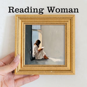 Pu&ograve; includere: Un dipinto incorniciato in oro intitolato "Reading Woman" raffigura una persona in abito bianco che legge vicino a una finestra. L'opera d'arte ha una tavolozza di colori tenui, con un focus sulla luce e l'ombra.