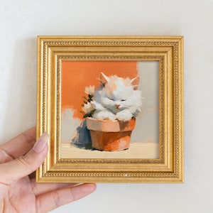 Op de afbeelding: Een klein vierkant schilderij in een sierlijke gouden lijst met een pluizig wit kitten met gesloten ogen dat rust in een terracotta-kleurige bloempot. De achtergrond toont een levendige oranje muur en een klein groepje witte bloemen.