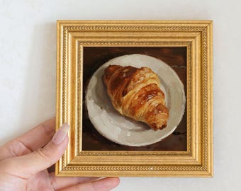 Könnte beinhalten: Ein kleines, gerahmtes Gemälde, das ein Croissant auf einem weißen Teller zeigt. Das Croissant ist goldbraun mit flockigen Schichten, der Teller ist cremeweiß. Das Bild wird von einer Hand gehalten.