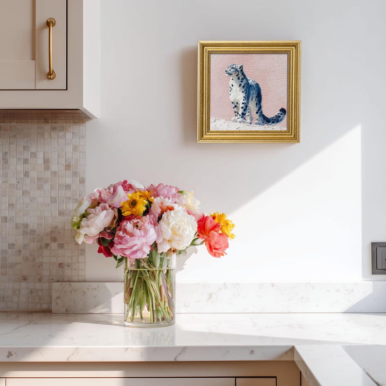 May include: A framed painting of a snow leopard with blue spots on a pink background hangs above a bouquet of colorful flowers in a clear vase. The flowers include pink, yellow, and orange blooms.