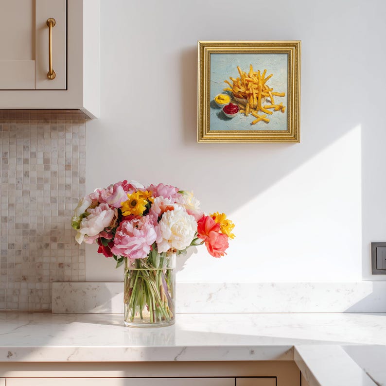 May include: A framed painting of chips with ketchup and mustard. The painting has a gold frame and hangs on a white wall. A bouquet of pink, yellow, and white flowers in a glass vase sits on a white countertop.