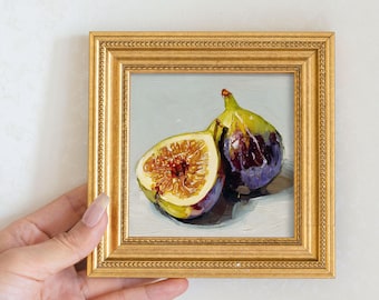 May include: A small, framed oil painting of two figs. One fig is whole, with a green and purple exterior. The other fig is cut open, revealing a detailed interior. The frame is gold and ornate.