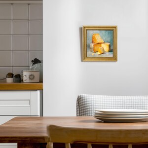 Peut inclure: Peinture encadr&eacute;e repr&eacute;sentant une nature morte de fromage. L'&oelig;uvre, dans un cadre dor&eacute;, pr&eacute;sente des morceaux de fromage jaunes et orang&eacute;s sur fond bleu clair. Le tableau est accroch&eacute; sur un mur blanc, mettant en valeur les couleurs.