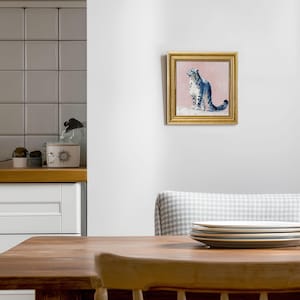 May include: A framed painting of a snow leopard. The artwork features a blue and white snow leopard on a pink background, set within a gold frame. The painting is displayed on a white wall.