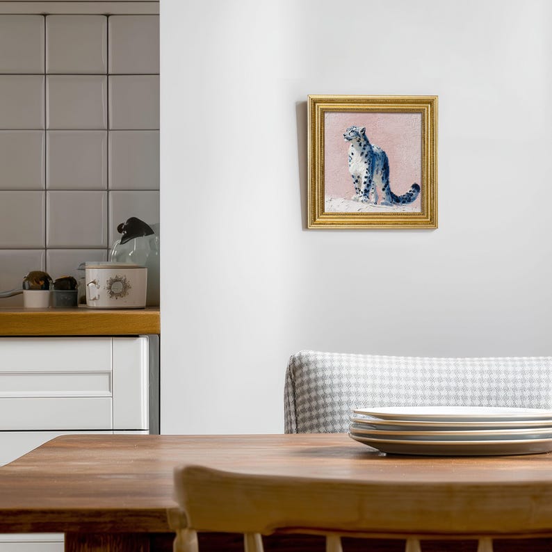 May include: A framed painting of a snow leopard. The artwork features a blue and white snow leopard on a pink background, set within a gold frame. The painting is displayed on a white wall.