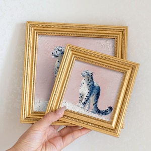 May include: Two framed paintings featuring snow leopards. The paintings have a pink background and gold frames. The snow leopards are depicted with blue and white spots. The frames are held by a person.