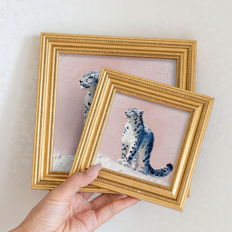 May include: Two framed paintings featuring snow leopards. The paintings have a pink background and gold frames. The snow leopards are depicted with blue and white spots. The frames are held by a person.