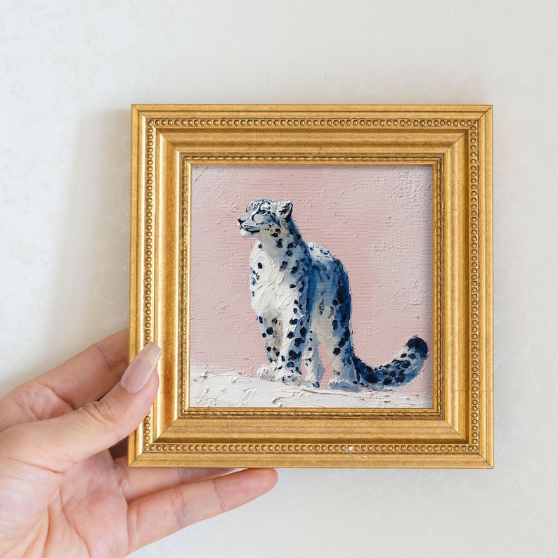 May include: A framed painting of a snow leopard with blue and white fur and black spots, set against a pink background. The artwork is in a gold-colored ornate frame. The painting is held by a hand.