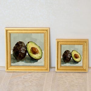 Op de afbeelding: Twee ingelijste avocadokunstwerken. E&eacute;n schilderij toont een hele avocado naast een gehalveerde avocado, waarbij de pit zichtbaar is. Het tweede schilderij toont een hele en een halve avocado. Beide schilderijen hebben gouden lijsten.