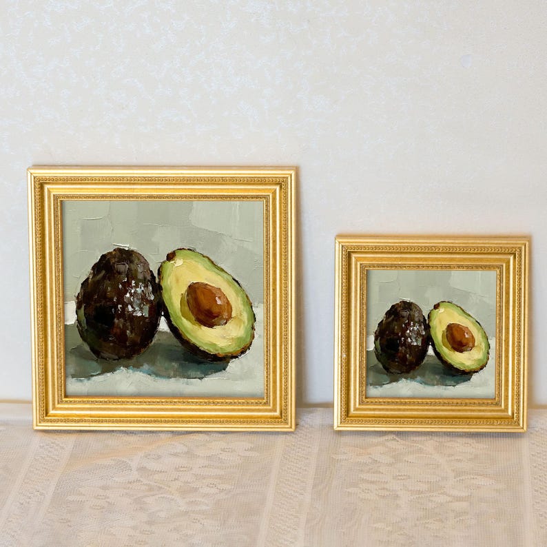 Op de afbeelding: Twee ingelijste avocadokunstwerken. E&eacute;n schilderij toont een hele avocado naast een gehalveerde avocado, waarbij de pit zichtbaar is. Het tweede schilderij toont een hele en een halve avocado. Beide schilderijen hebben gouden lijsten.