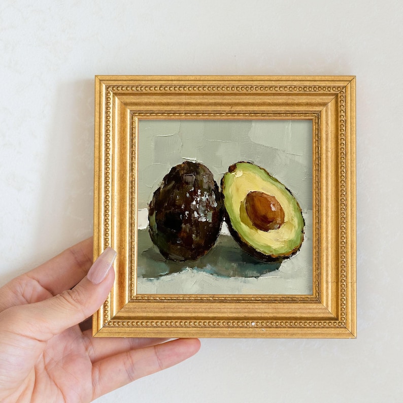 Op de afbeelding: Een ingelijst olieverfschilderij van twee avocado's, &eacute;&eacute;n heel en &eacute;&eacute;n doormidden gesneden, met een bruine pit. Het schilderij heeft een lichtgroene achtergrond en een gouden, sierlijke lijst. Het kunstwerk wordt door een persoon vastgehouden.