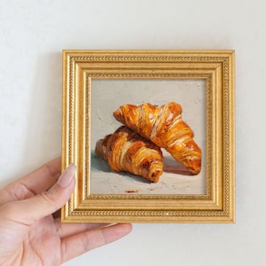 May include: A small, framed painting of two golden-brown croissants. The painting is in a gold frame with a detailed border. The croissants are painted with detailed brushstrokes, showing their flaky texture and rich colour. The background is a light grey.